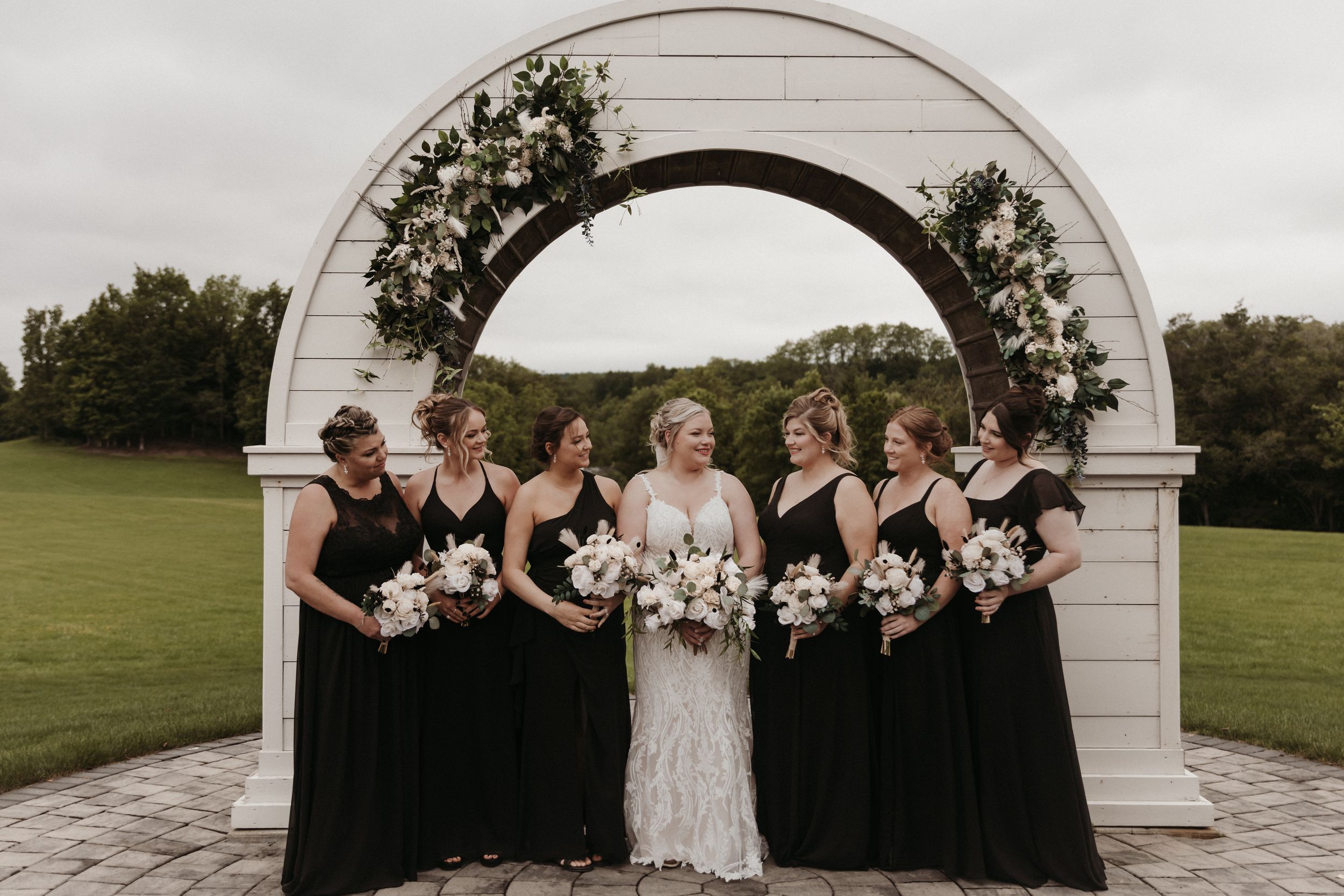A wedding party in black and white attire poses together under an elegant white archway at an outdoor venue.