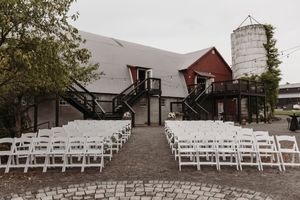 Rustic barn wedding venue with white chairs set up for outdoor ceremony and silo in background.