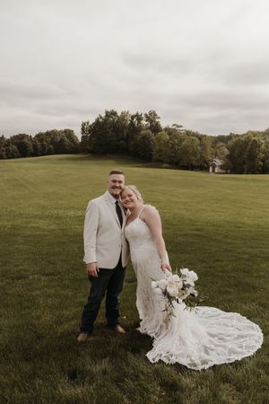 A couple poses in a grassy field during a wedding photoshoot with a beautiful cloudy sky backdrop.
