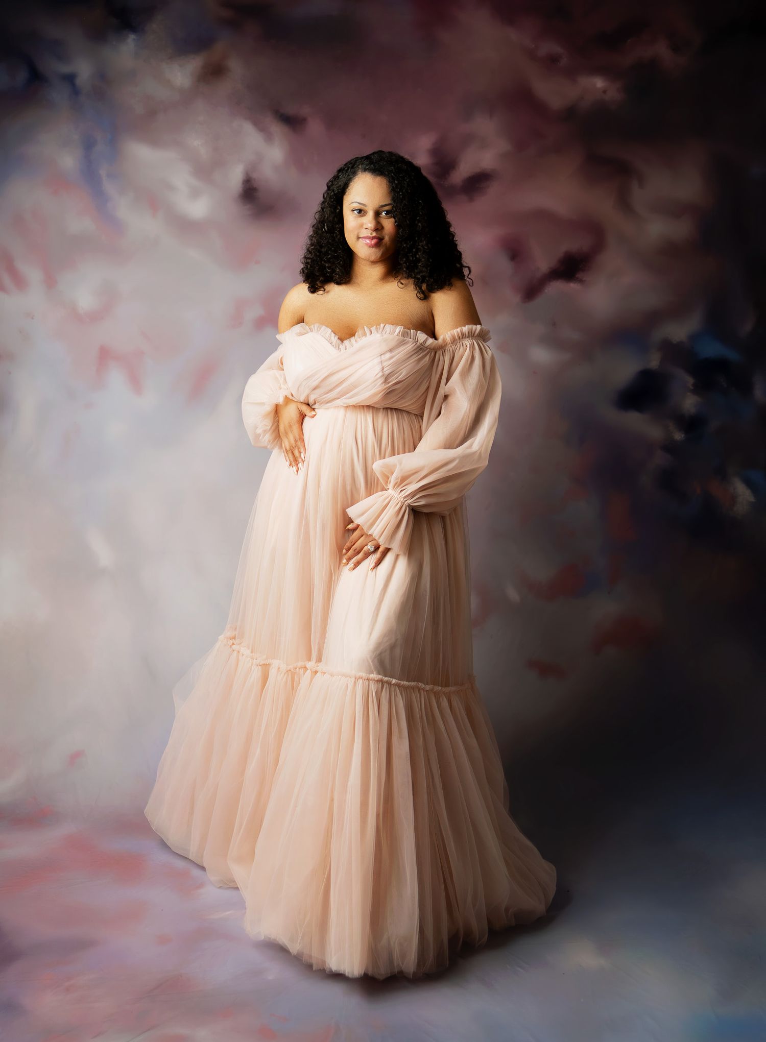 Ethereal maternity photoshoot featuring a flowing peach tulle gown against a moody clouded backdrop.