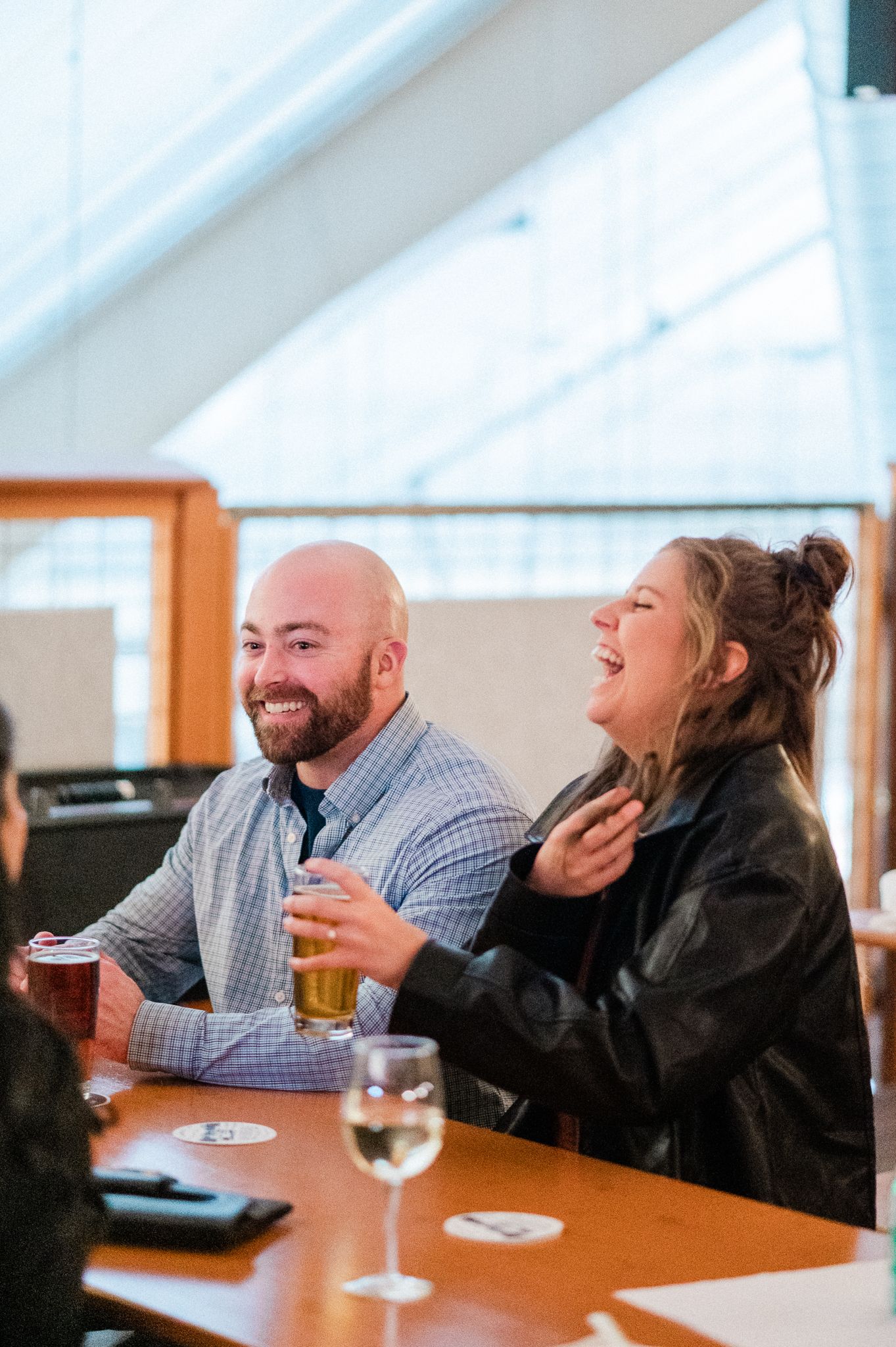 Two friends share a laugh while enjoying drinks at a wooden table in a bright, casual dining setting.
