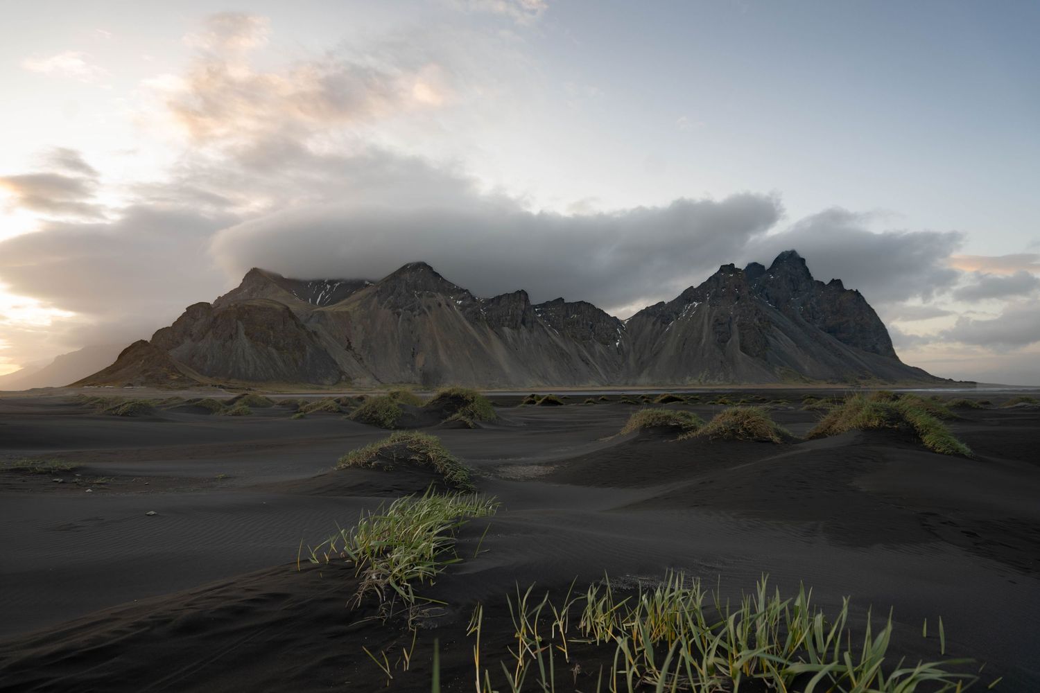 Jagged mountain peaks rise above black sand dunes with sparse grass at golden hour under dramatic clouds.