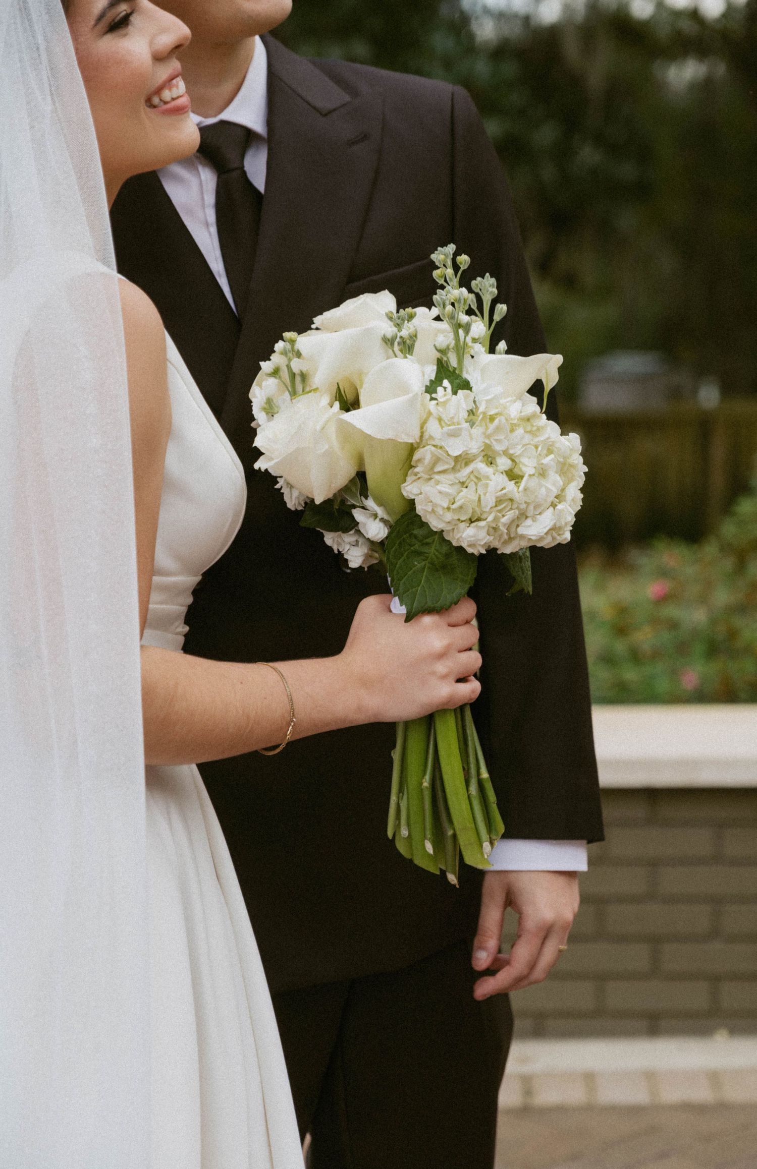 A bride in white holds an elegant bouquet of white calla lilies and roses while standing next to someone in a black suit.