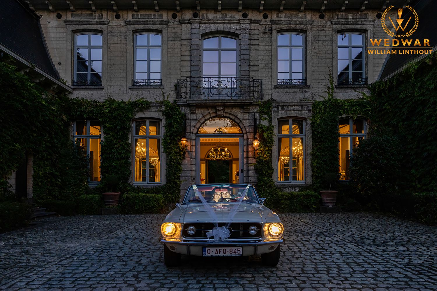 Newlywed couple poses romantically by vintage car in black and white wedding photograph.