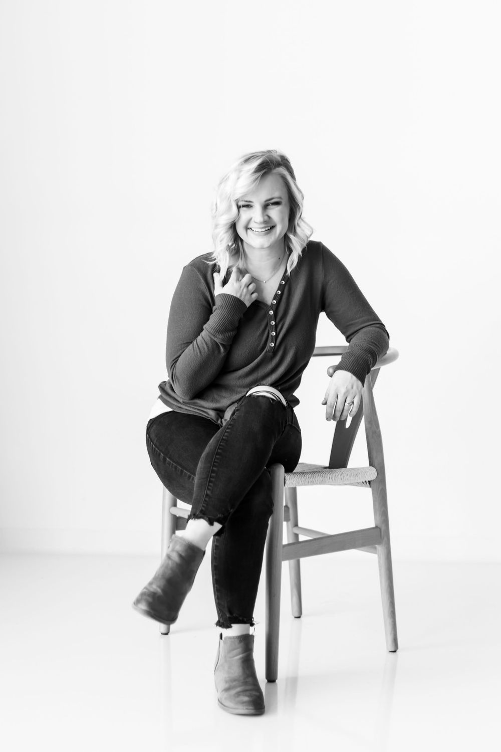 Black and white portrait of someone sitting casually on a white chair wearing a sweater, jeans and boots.