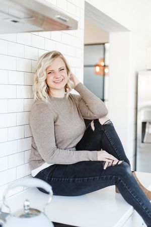 Woman in gray sweater and dark jeans poses casually on white countertop in bright modern kitchen setting.