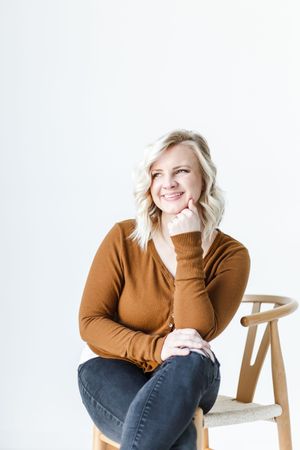 Person in brown sweater and jeans sits casually on wooden chair against white background in studio setting.