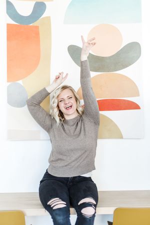Person in gray sweater and ripped jeans sitting against colorful geometric wall art showing joyful expression.