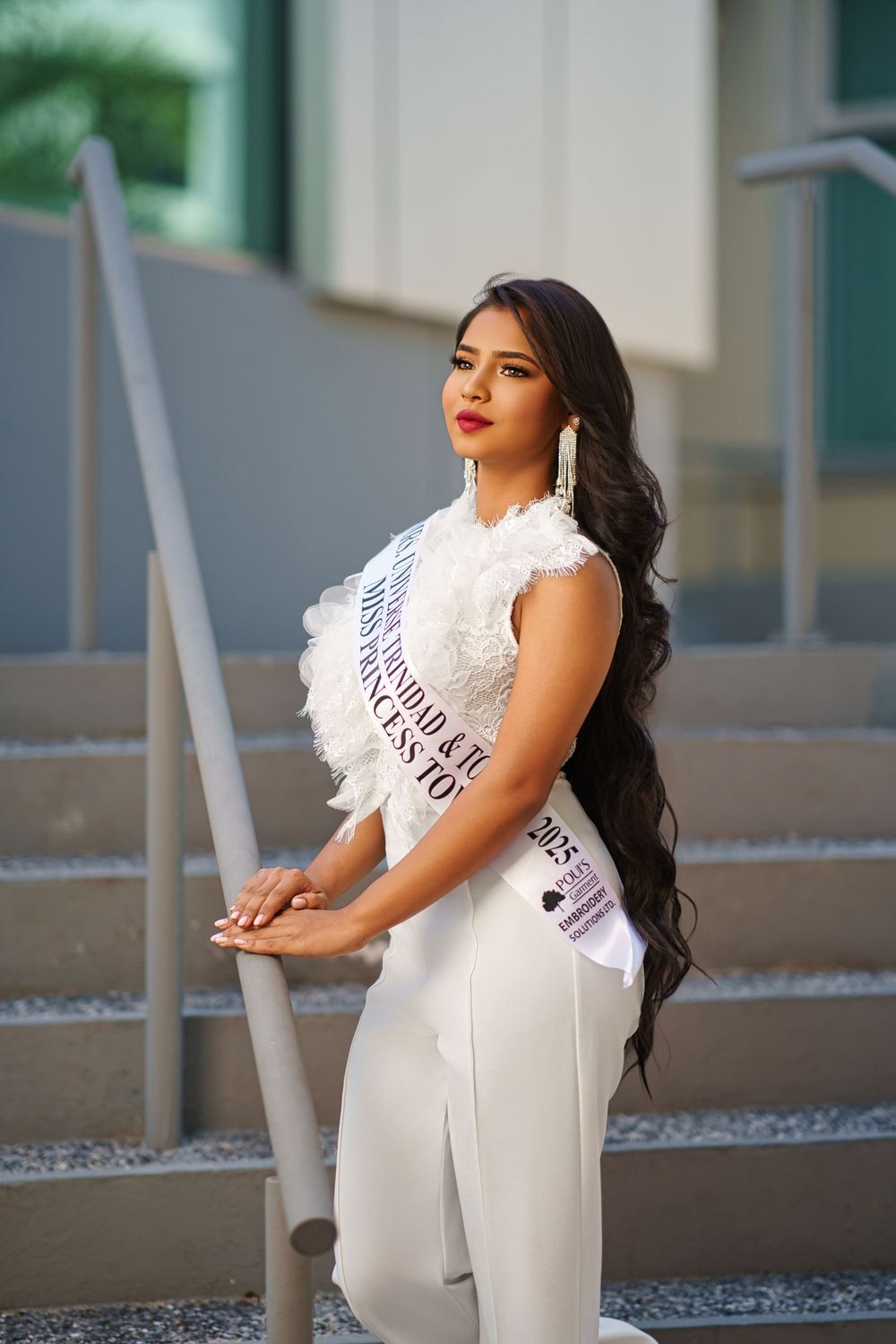 A pageant contestant in a white jumpsuit and sash poses elegantly on concrete steps.