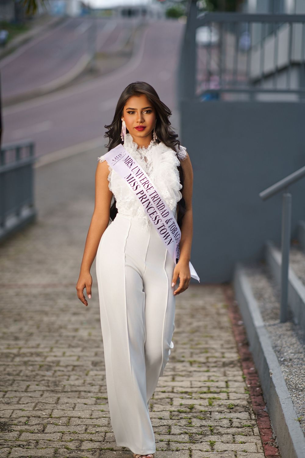 A beauty pageant contestant wearing an elegant white jumpsuit and sash stands on a paved walkway.