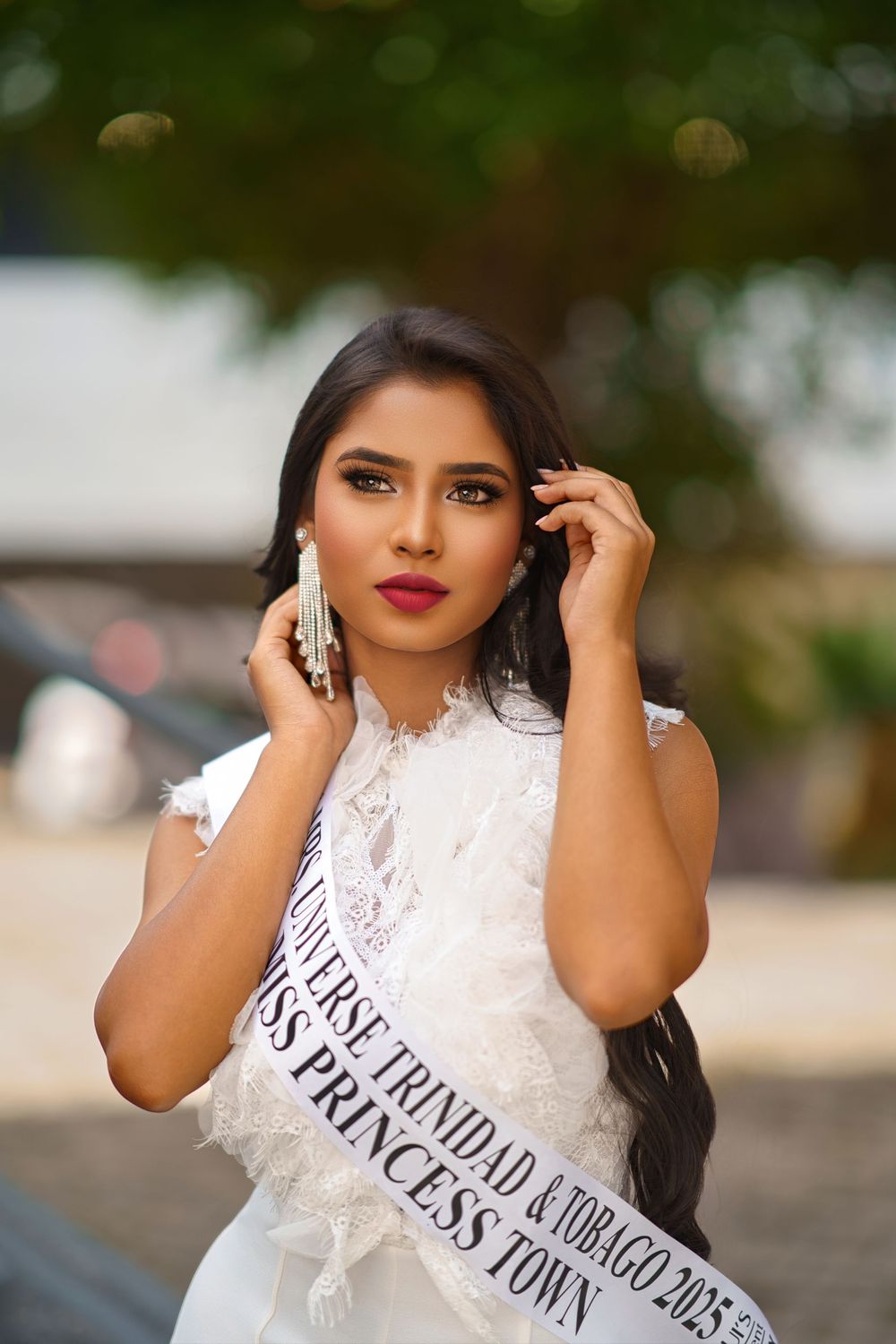 A beauty pageant contestant wearing a white lace dress and sash poses elegantly against a blurred outdoor background.