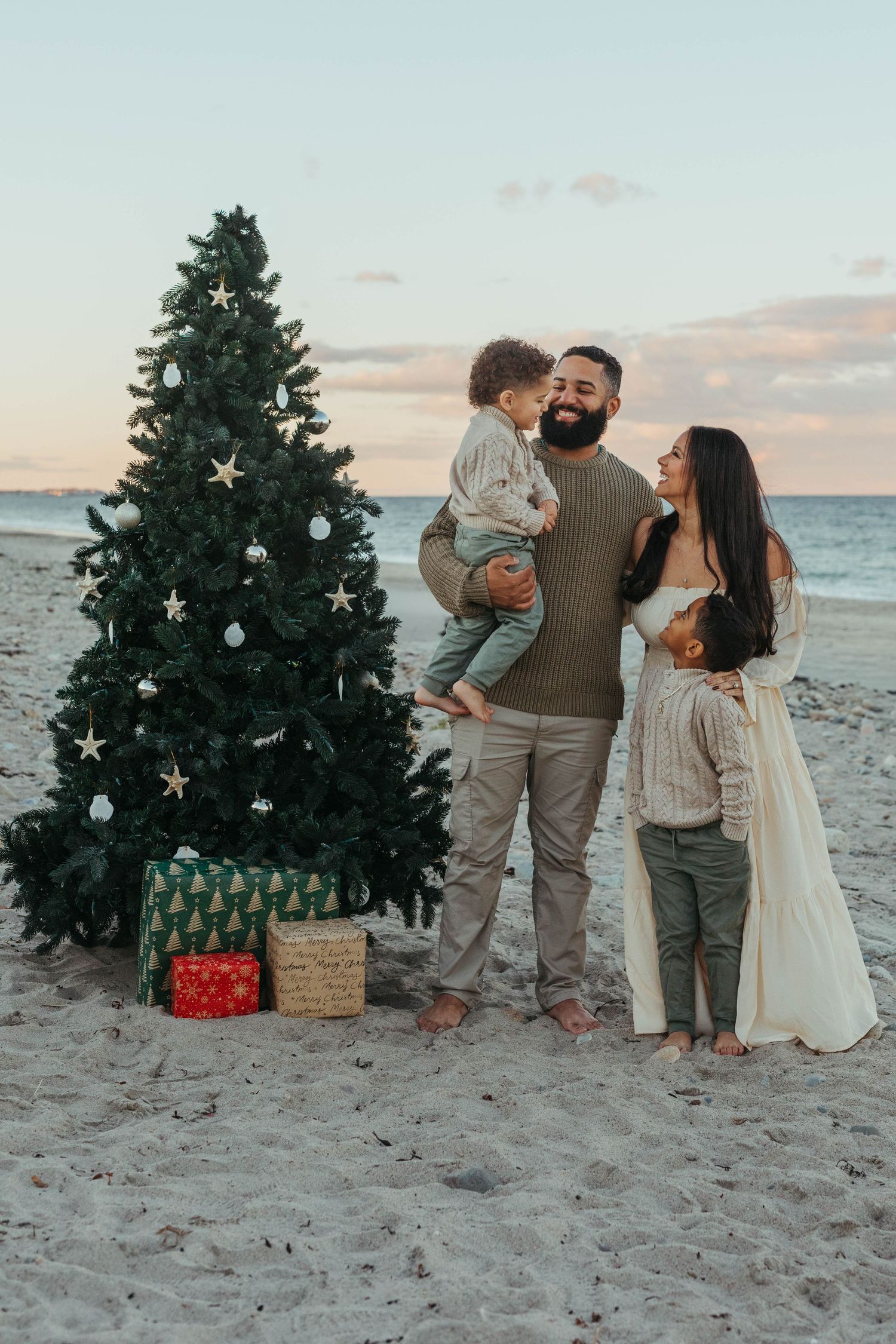 Family poses on beach at sunset with decorated Christmas tree creating a festive holiday scene.