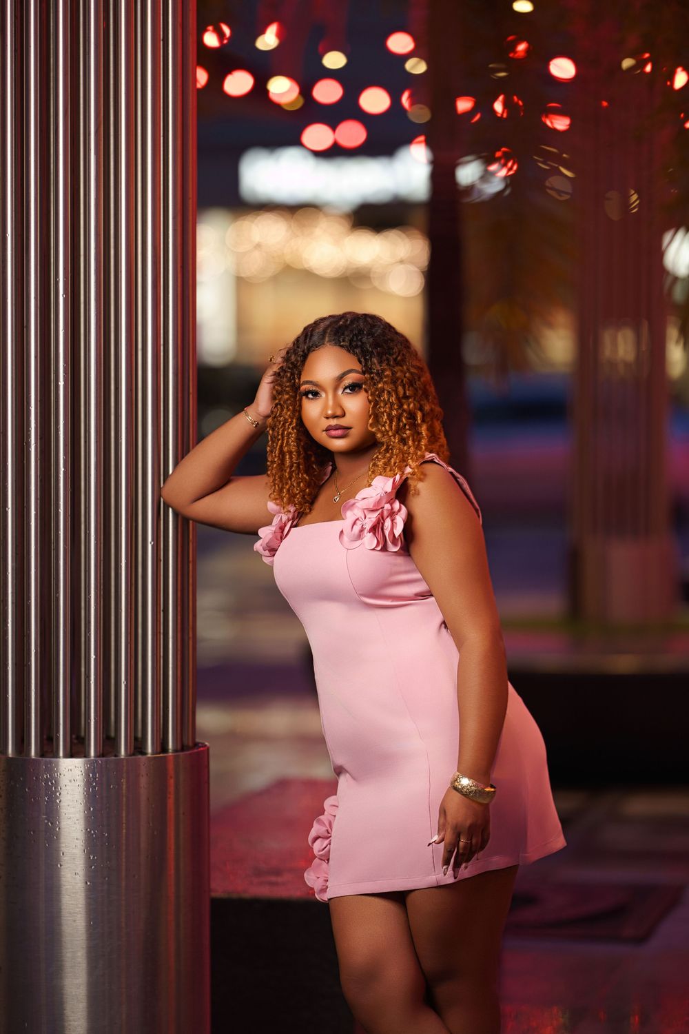 Evening portrait session with metallic columns and pink dress against glowing city lights in background.