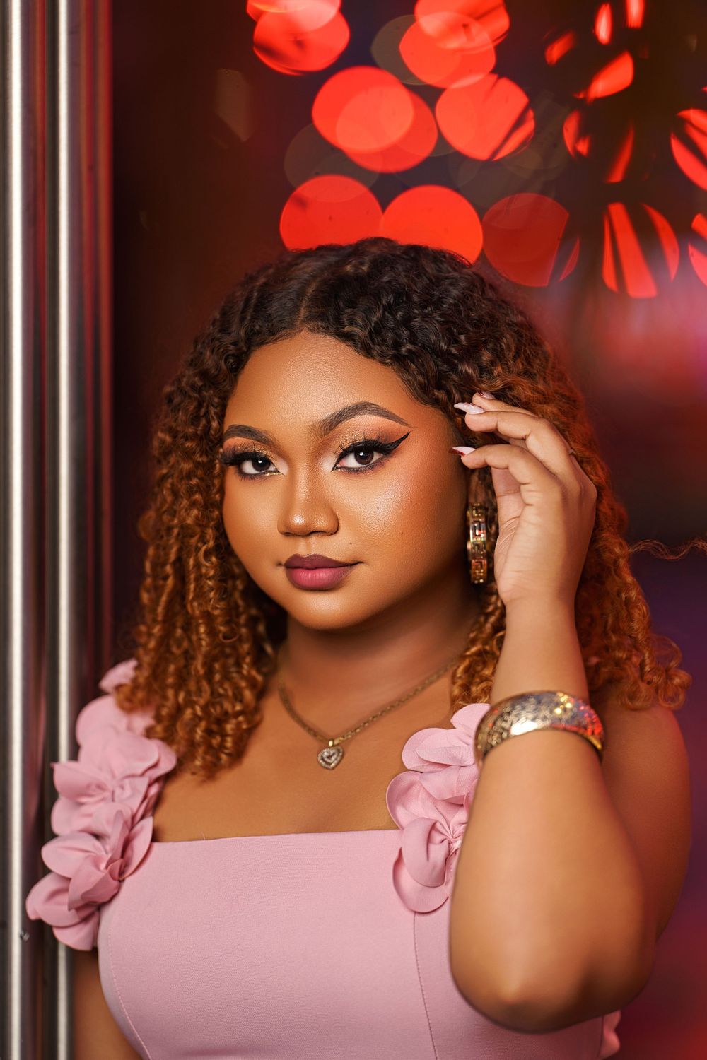 A woman in a pink dress poses confidently with curly hair and glamorous makeup against a bokeh background.