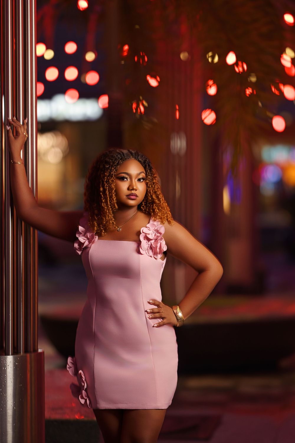 A stylish pink dress with floral details photographed against a nighttime cityscape with red lights.