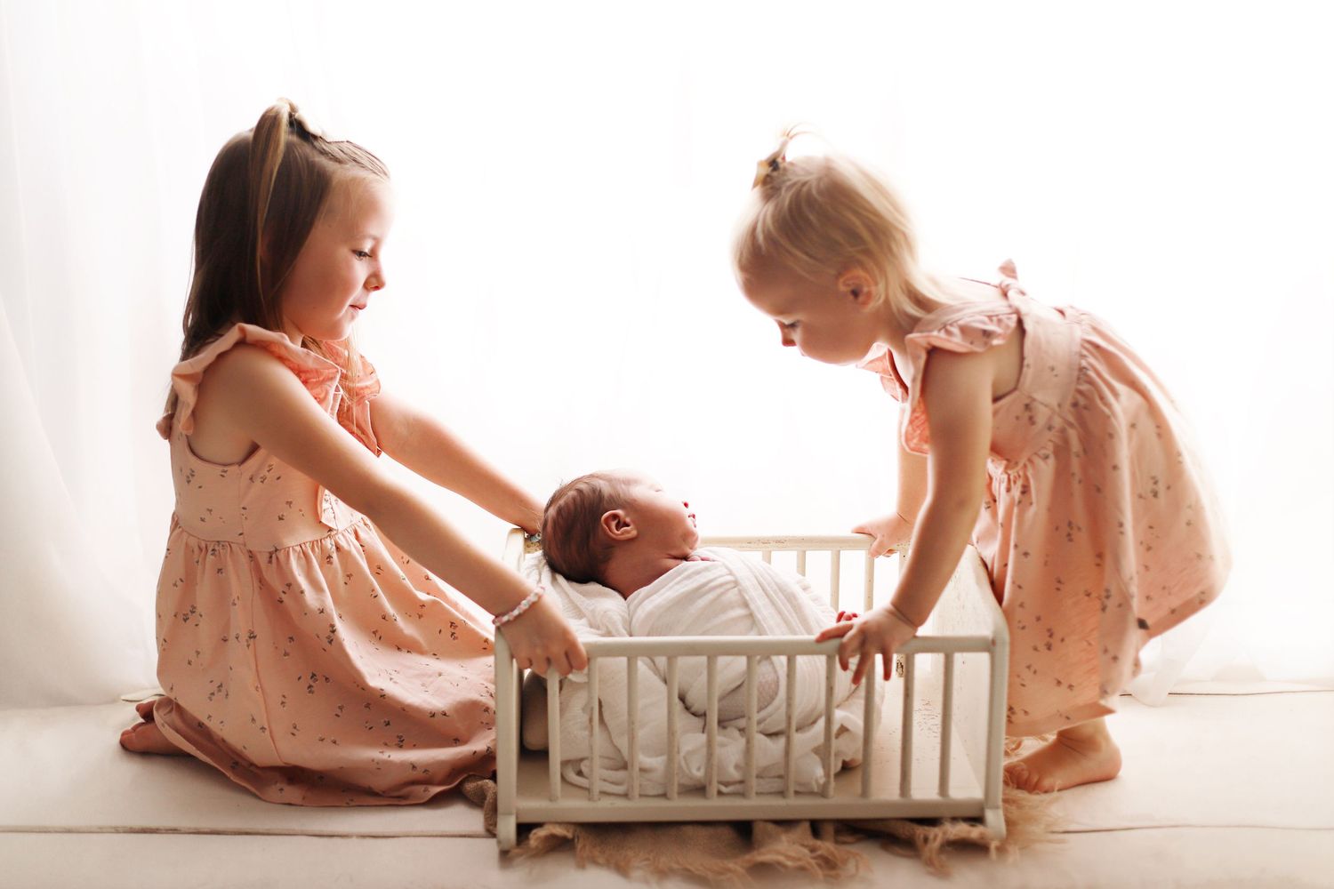 Two children in matching peach dresses interact with a white crib in a softly lit room.