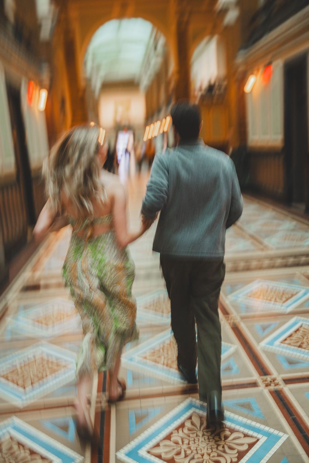 A couple runs down an ornate hallway with geometric patterned floors and warm lighting creating a romantic ambiance.