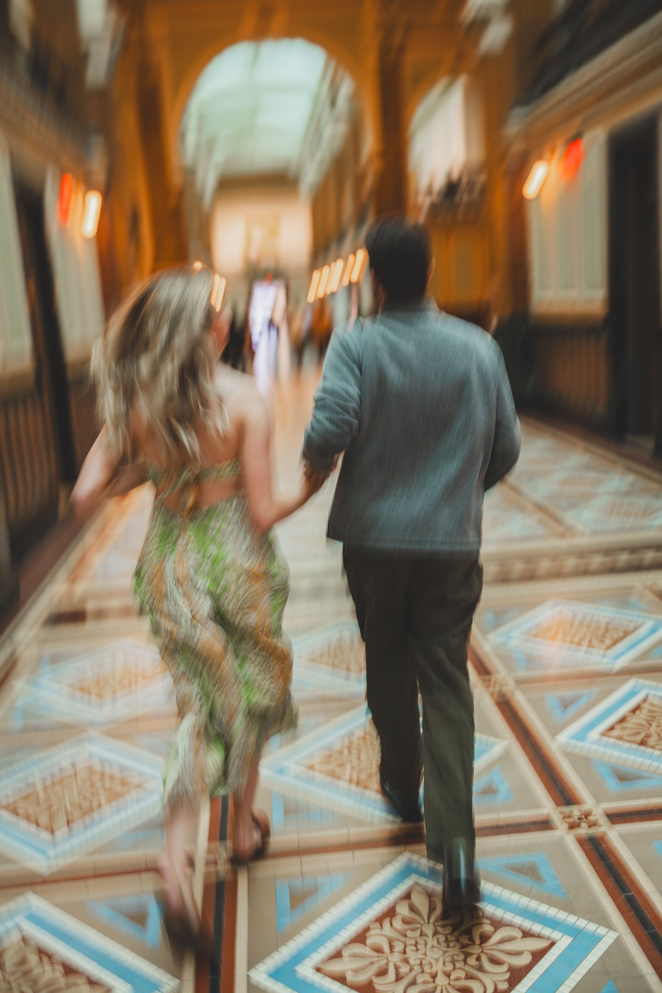 A couple runs down an ornate hallway with geometric patterned floors and warm lighting creating a romantic ambiance.