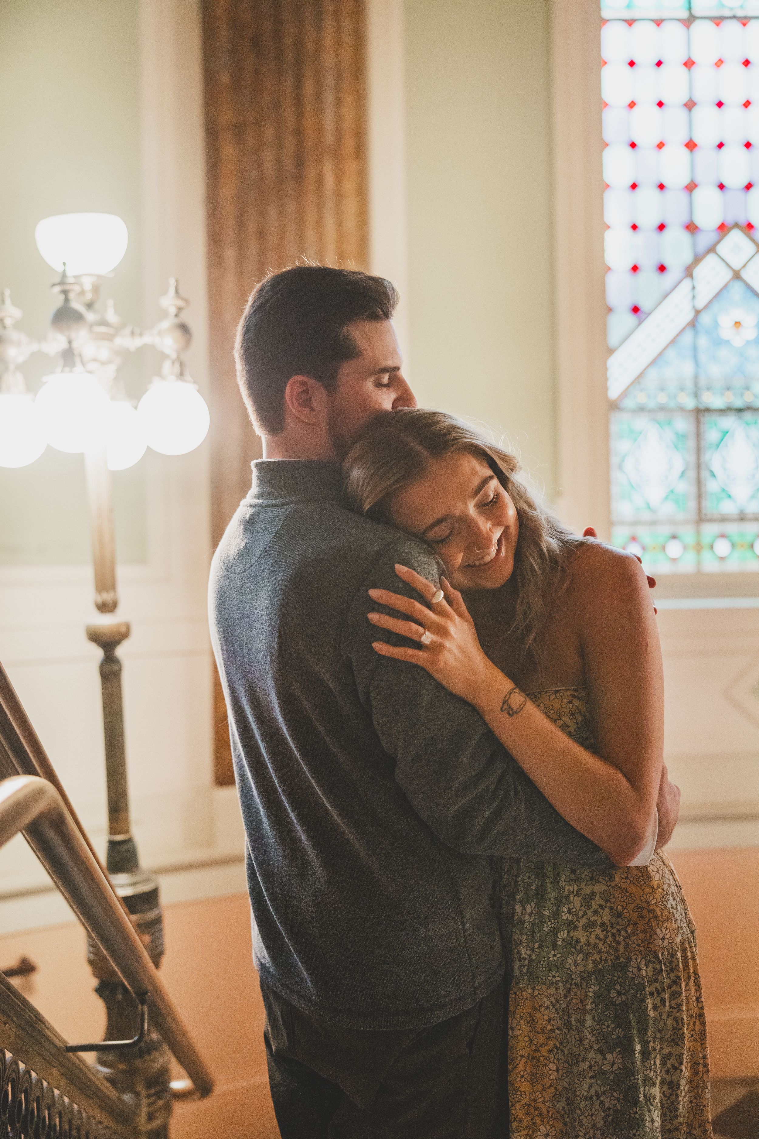 A romantic couple embraces in a warm glow by a stained glass window in an elegant interior setting.