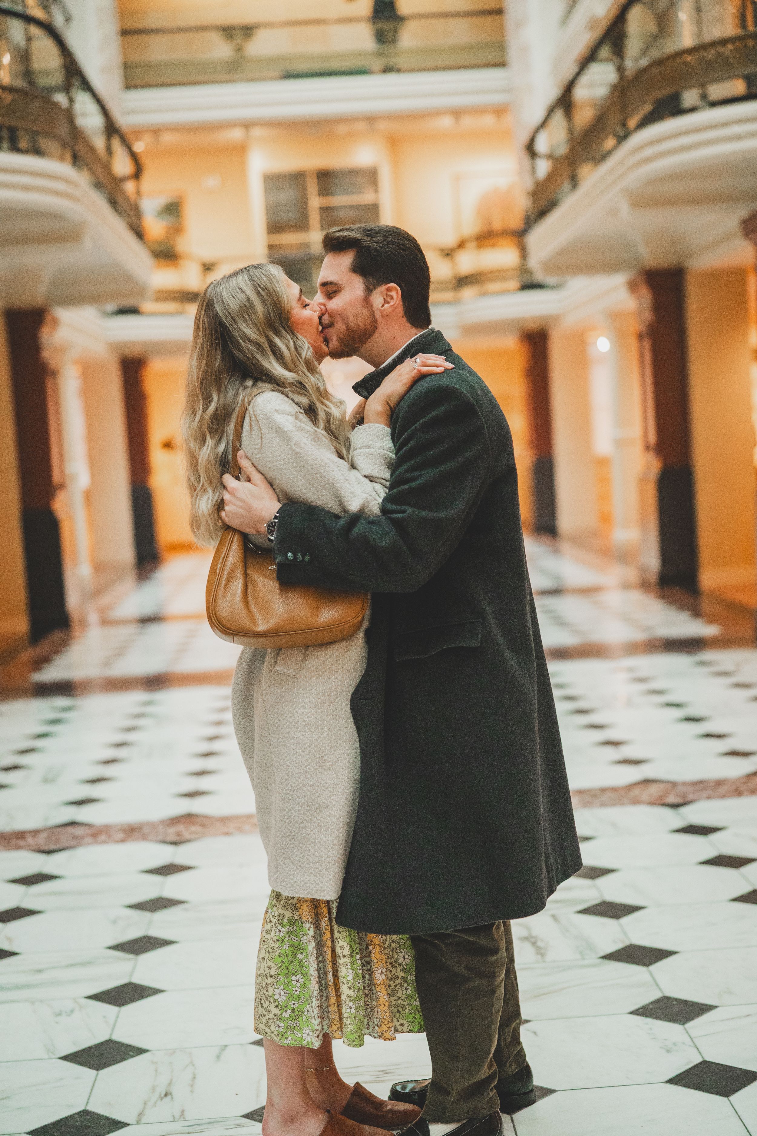 A romantic couple embraces and dances on a checkered floor in an elegant hallway with peach-colored walls.