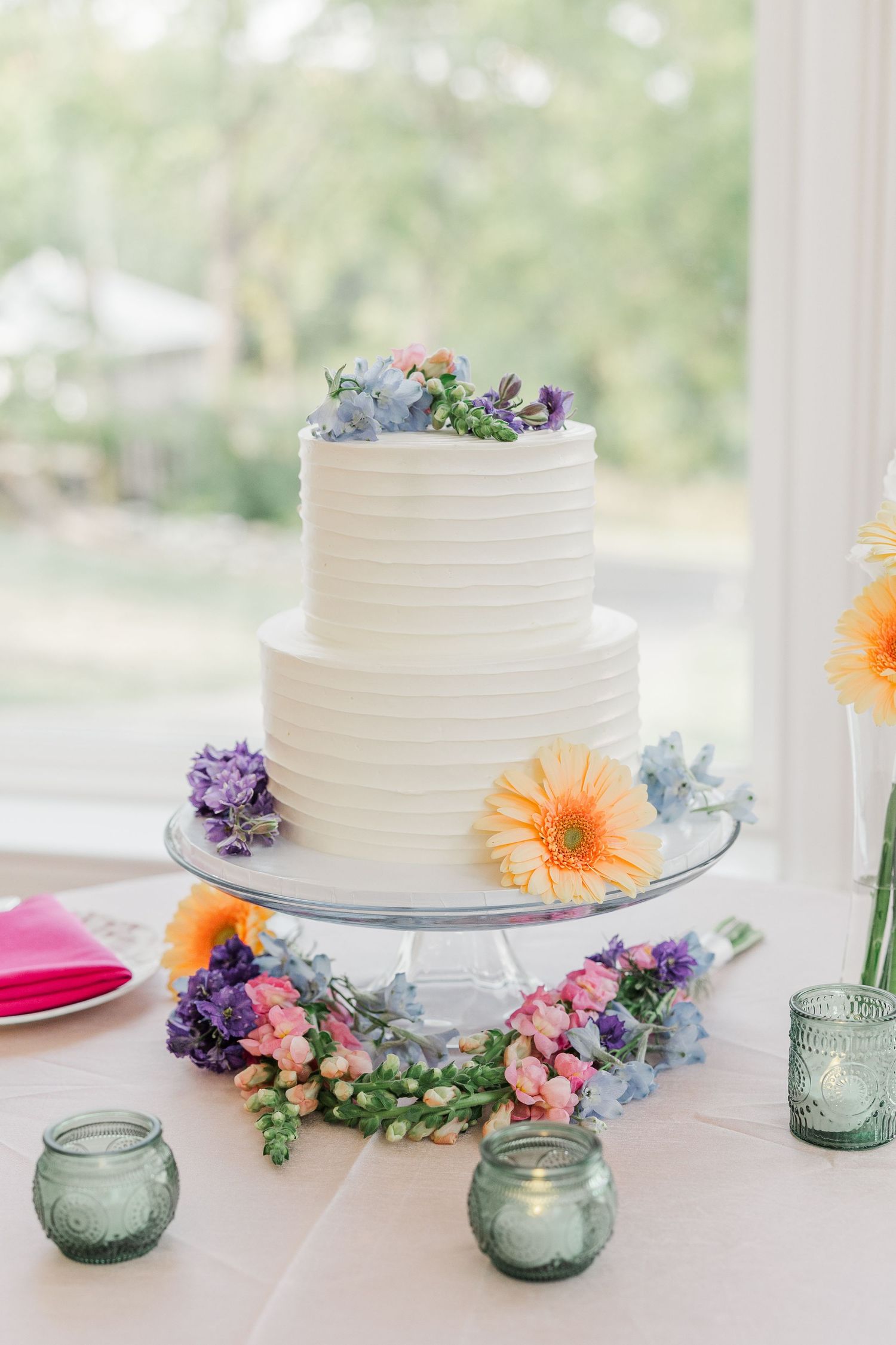 White wedding cake decorated with fresh flowers and surrounded by blue glass votives on an elegant table setting.