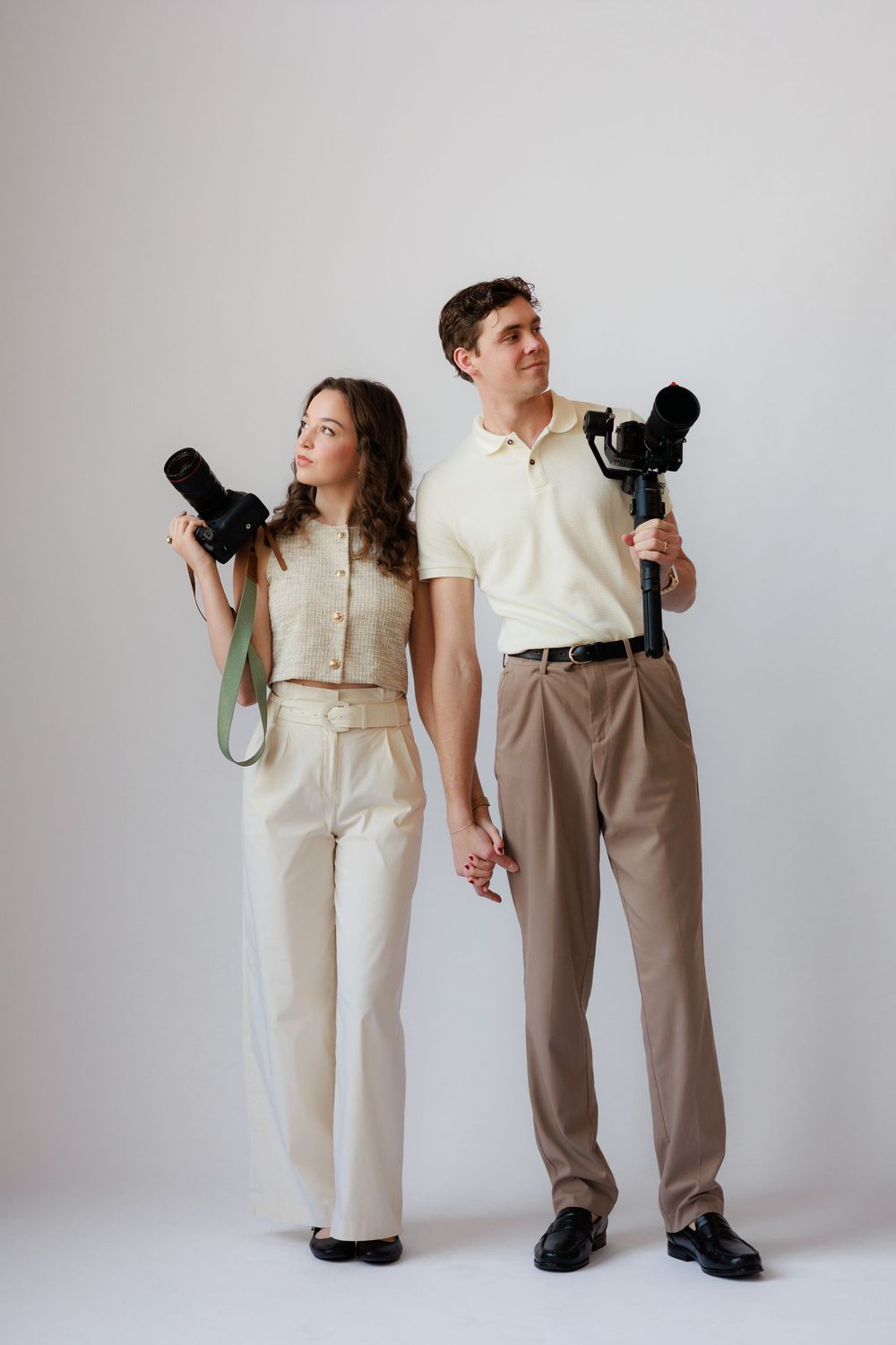 A couple in vintage-style clothing poses playfully with cameras against a white backdrop in a series of artistic shots.