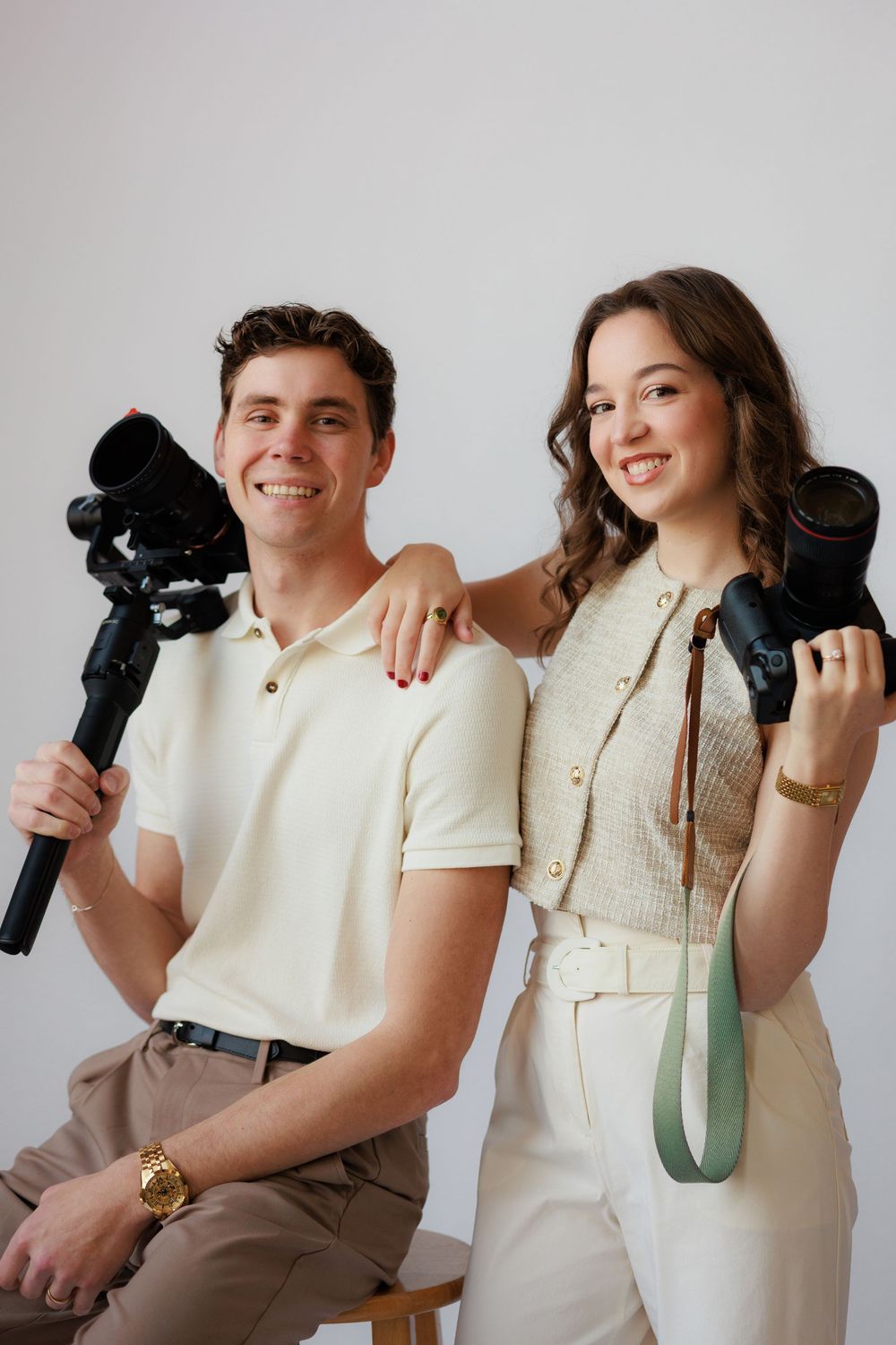 Two photographers pose with professional camera equipment while wearing casual cream and white outfits.