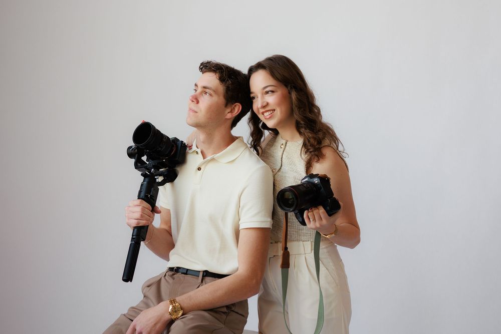 Two photographers stand back to back holding professional DSLR cameras against a white background.