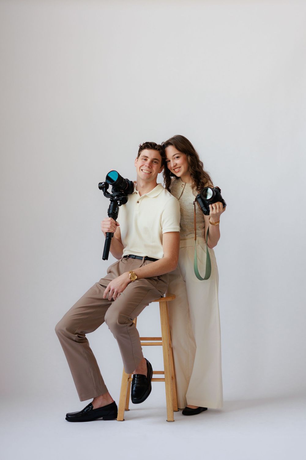 Two photographers in neutral tones pose together with cameras against a white studio backdrop.