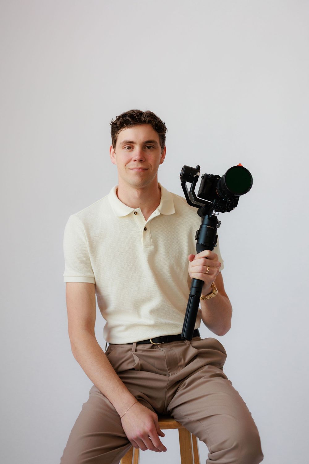 A photographer in a white polo and khaki pants poses with a camera against a light gray backdrop.