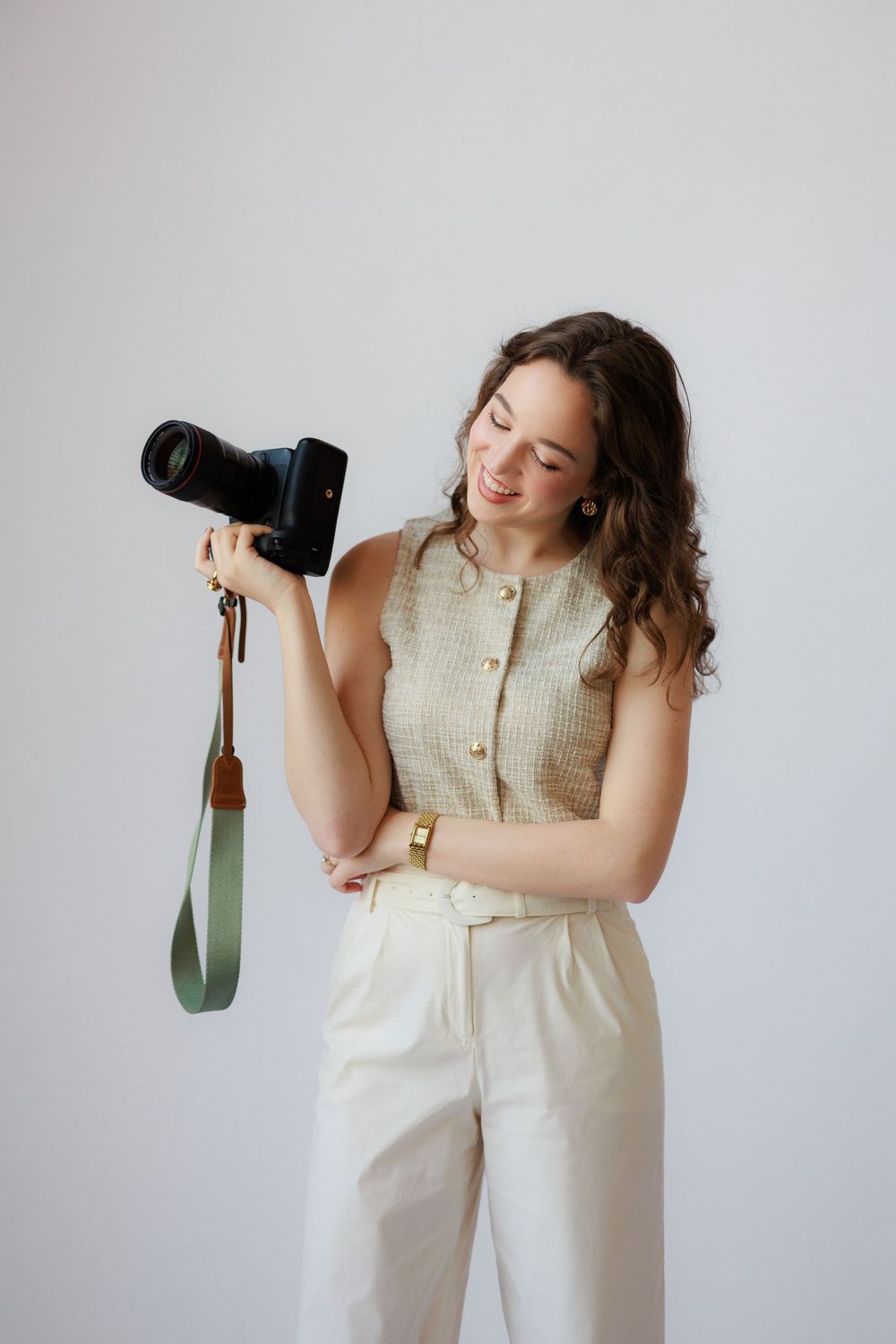 A photographer in a cream colored outfit poses with a professional camera against a white background.
