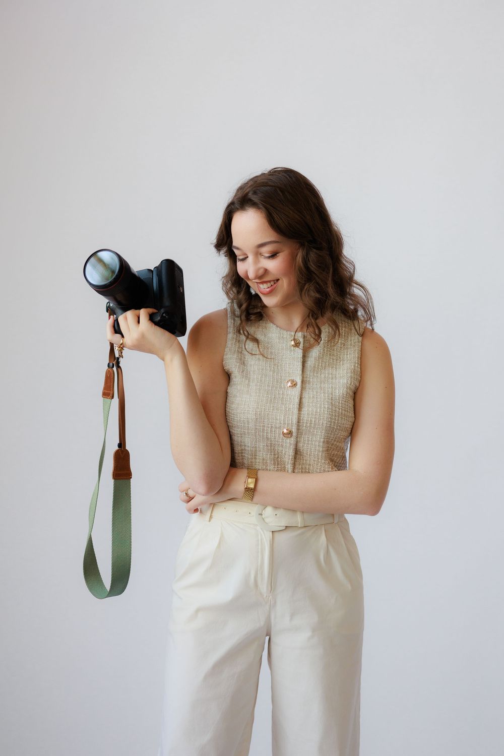 A photographer in a cream colored outfit holds a camera against a white studio backdrop.