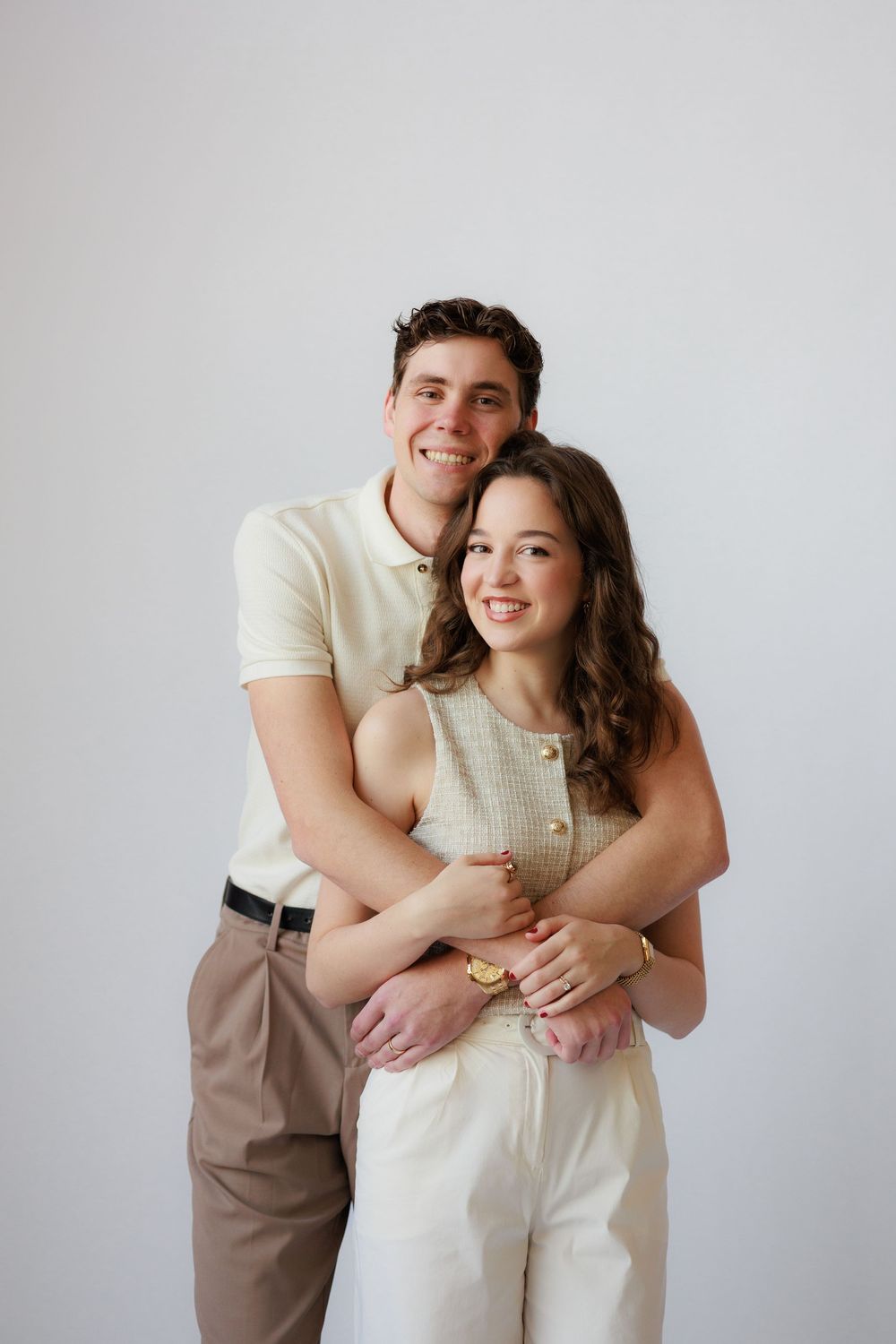 A couple in neutral-toned clothing embraces and smiles against a light gray studio backdrop.