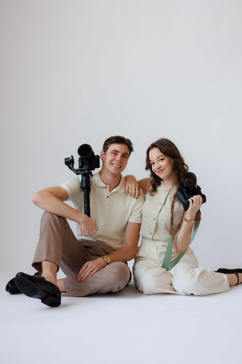 Two photographers laying on the floor with cameras in a minimalist white studio setting.