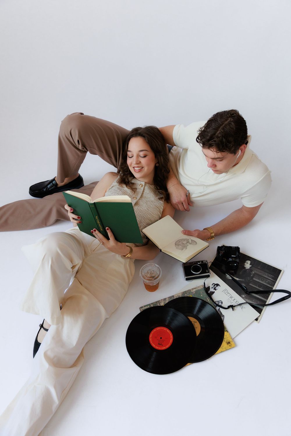 Two people lying down and reading books together while vinyl records sit nearby on a white background.