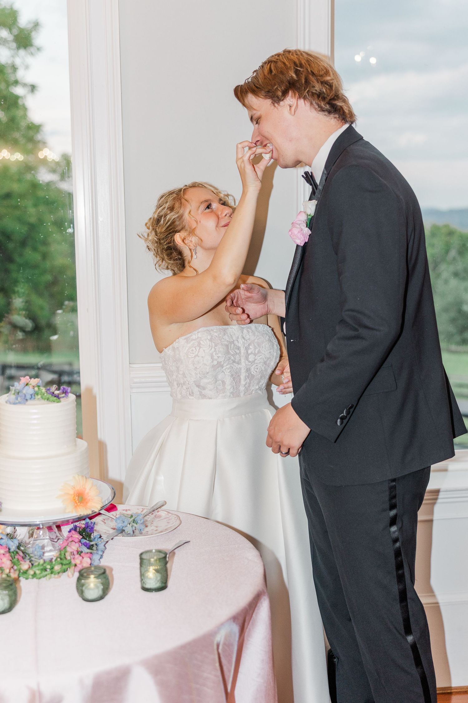 A wedding couple sharing cake during their reception beside a decorated dessert table.