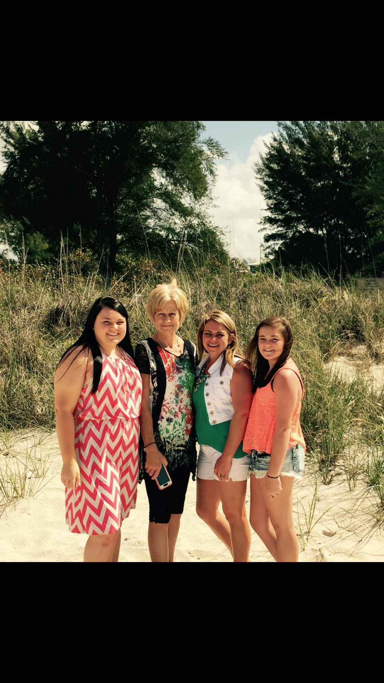 Four friends in swimsuits pose together on a sunny beach with sand dunes and trees in the background.
