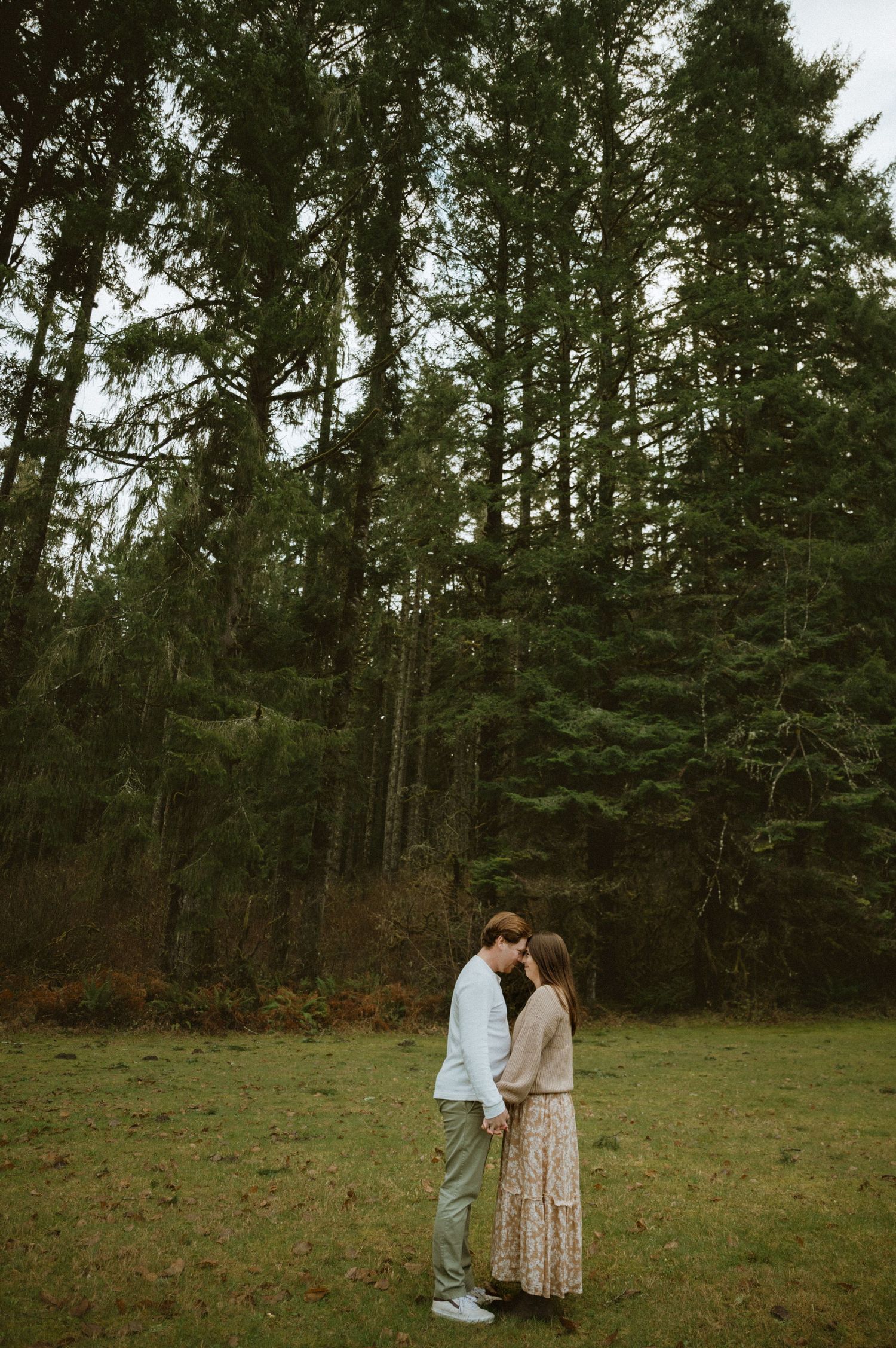 A couple shares a tender moment in a serene forest clearing surrounded by tall evergreen trees.