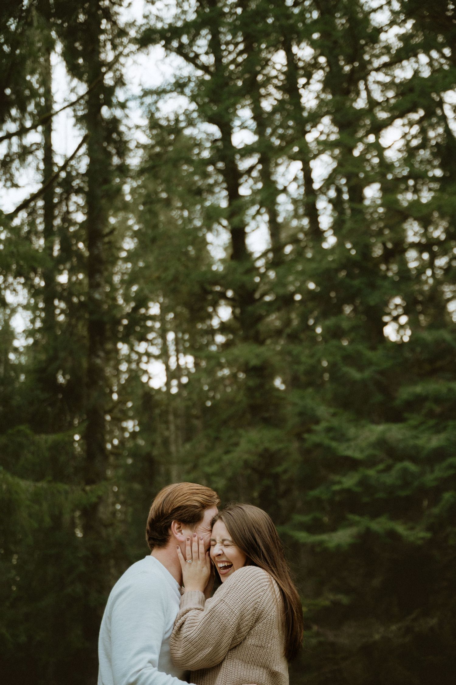 A romantic couple shares an intimate moment while laughing together in a lush evergreen forest setting.