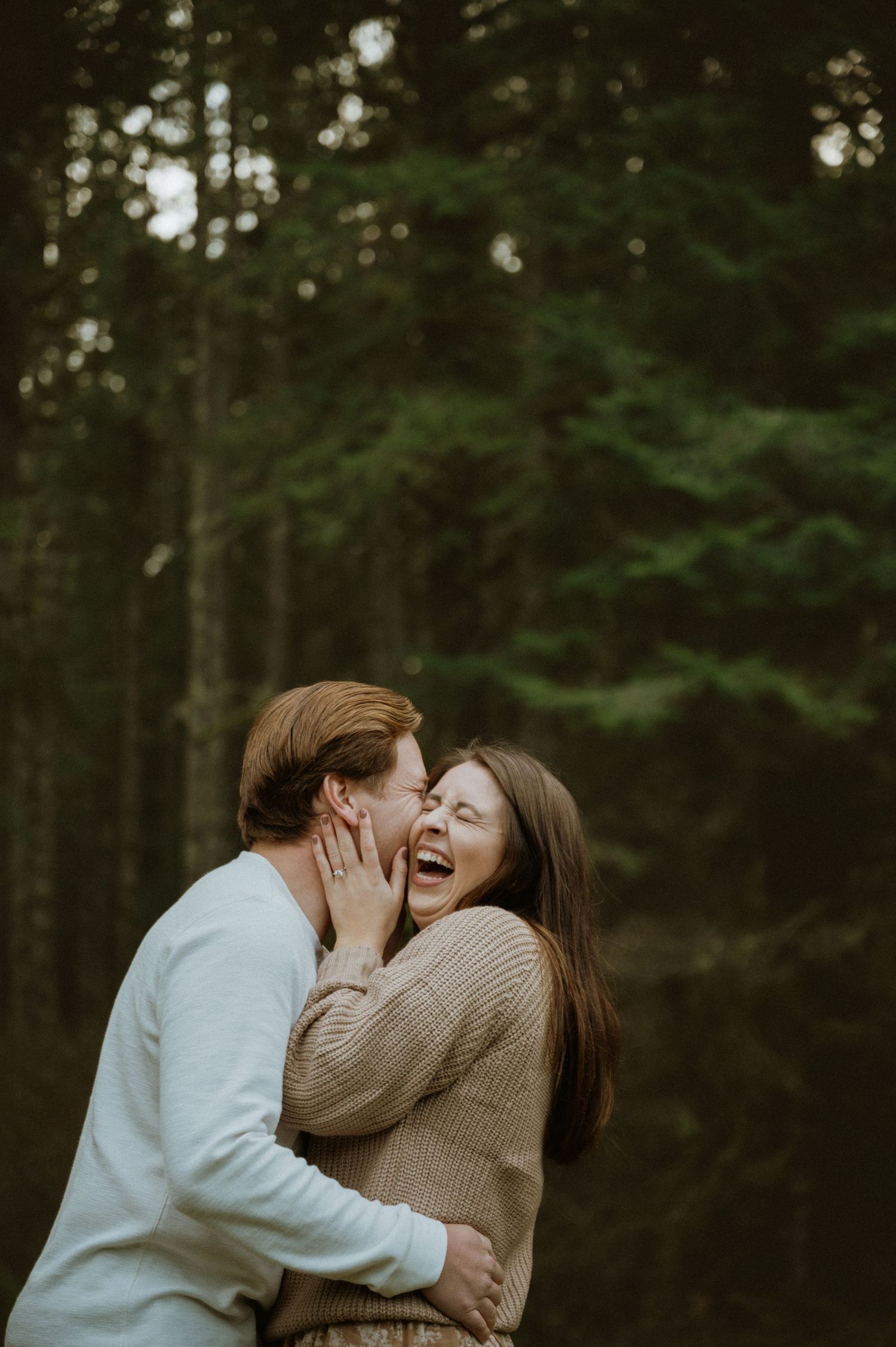 A couple shares an intimate embrace while laughing together in a forest setting during golden hour.