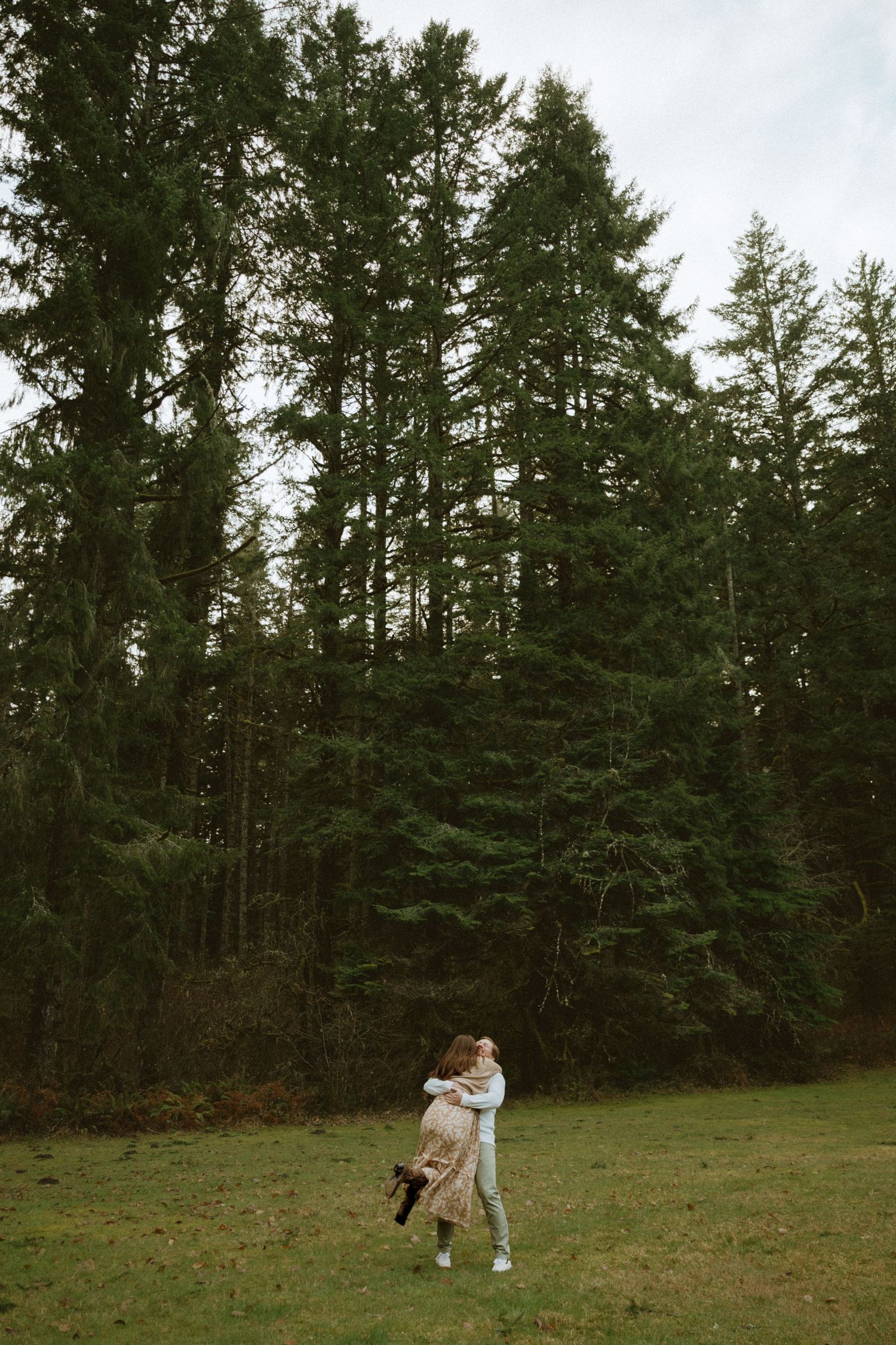 Two people dance playfully in a grassy meadow surrounded by tall evergreen trees on an overcast day.