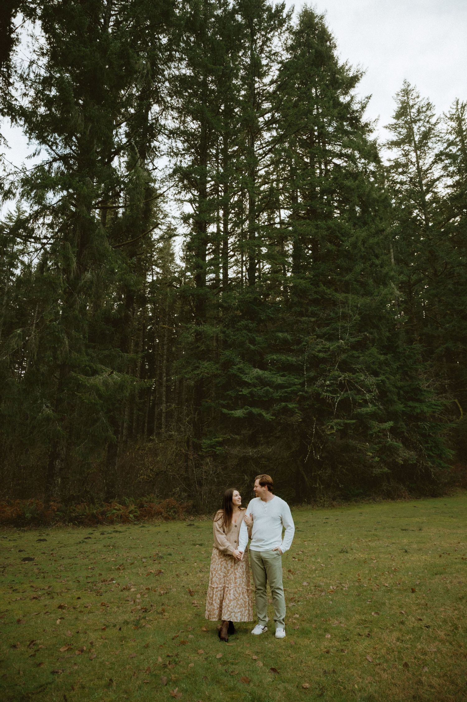 A couple stands together on a grassy field with tall evergreen trees creating a dramatic forest backdrop.