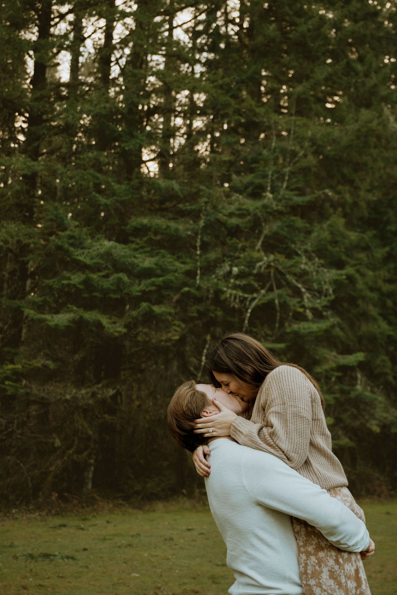Two people in light colored sweaters share an intimate moment against a backdrop of tall evergreen trees.