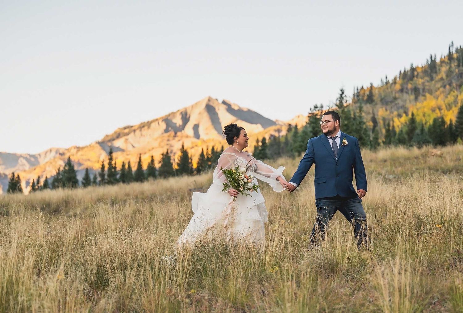 Wedding couple walks through golden mountain meadow at sunset as bride's dress and veil flow in the wind.