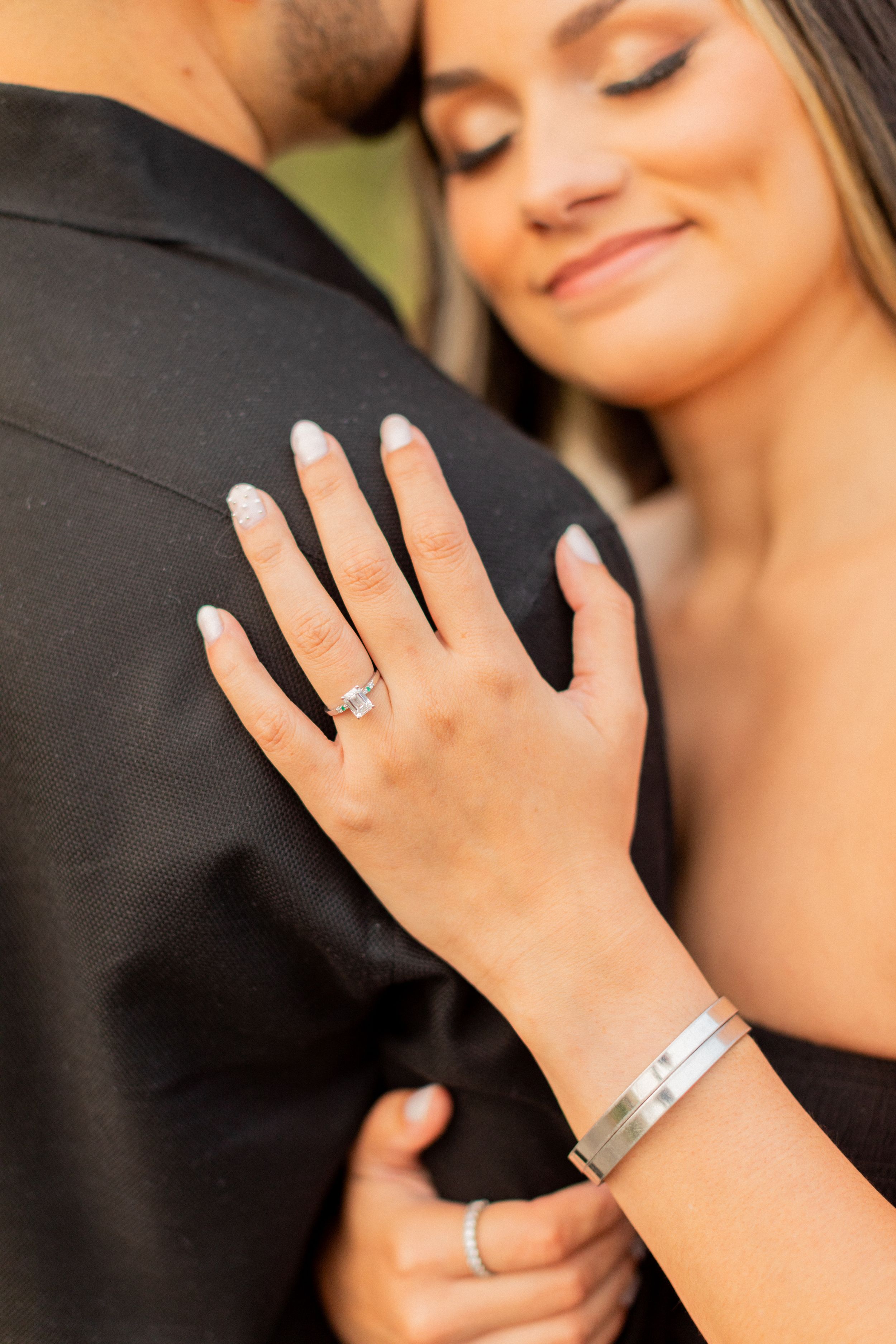 Close up of engagement ring and silver bracelet as couple embraces.