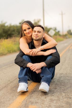 A couple sits intimately on a rural road with yellow lines, sharing an embrace in casual attire.