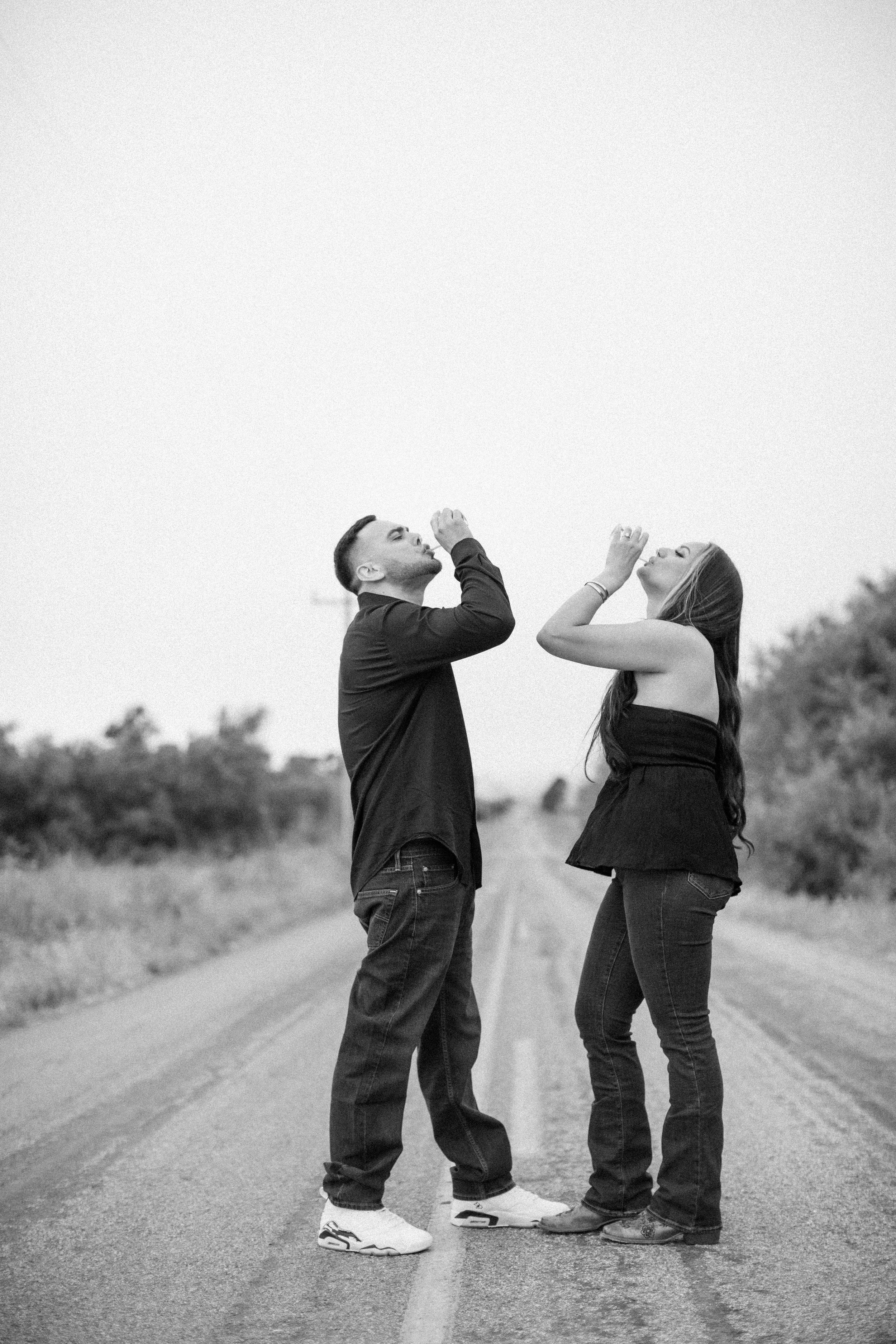 A playful couple stands on a dirt road taking silly photos in black and white.