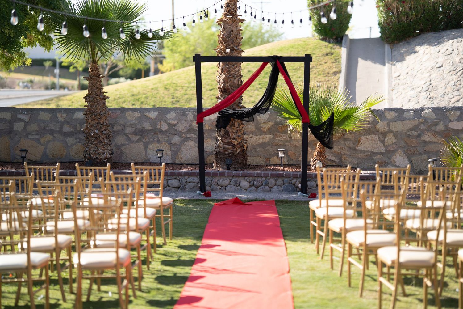 Wooden chairs line up in rows for an outdoor garden wedding ceremony with a red carpet aisle and palm trees.