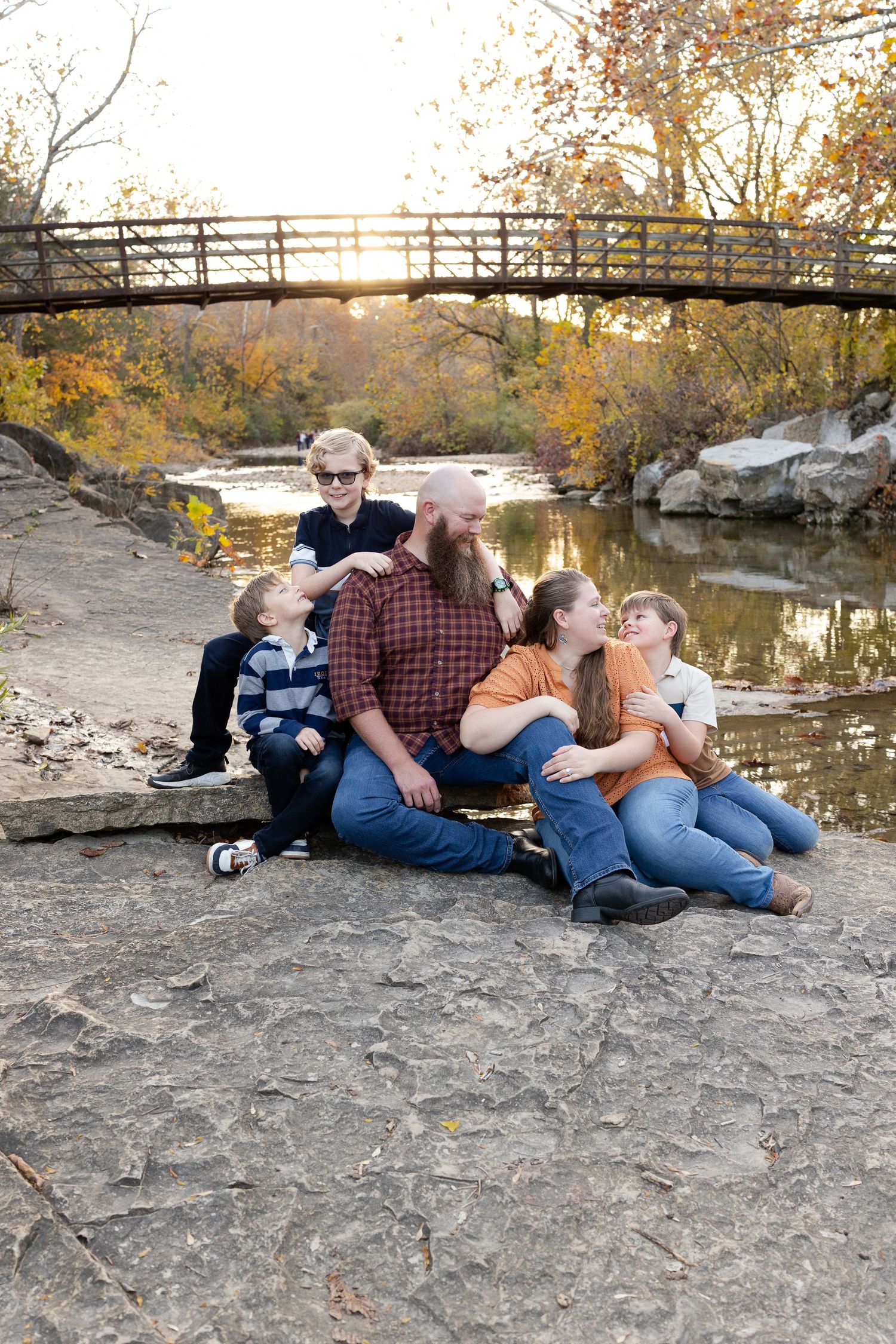 Family relaxing together on rocky riverbank during autumn sunset with bridge in background.