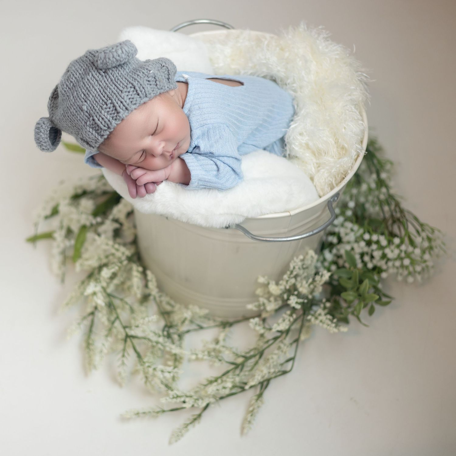 Newborn baby in gray hat sleeps peacefully in white bowl surrounded by delicate white flowers and greenery.